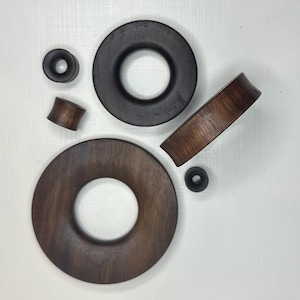 May include: A selection of dark brown and black wooden ear plugs and gauges. The image shows a variety of sizes and styles, including circular and cylindrical designs. The wood grain is visible on some pieces, enhancing their natural appearance.