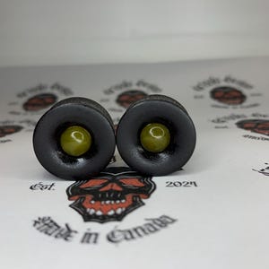 May include: Two black ear gauges with olive green centers. The gauges are circular with a flared design. The background features a skull logo and the text "Made in Canada."