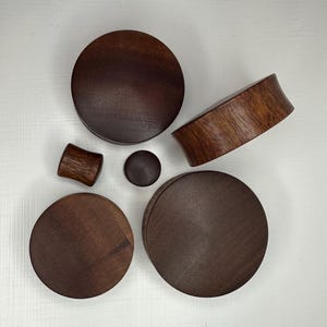 May include: Five wooden ear gauges in various sizes, ranging from small to large. The gauges are all a dark brown colour and have a smooth, polished finish.