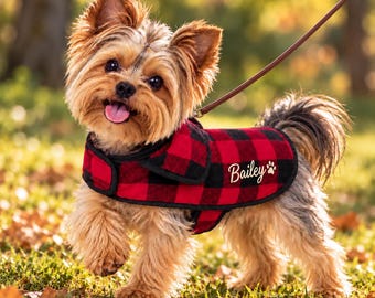 Può includere: Un piccolo Yorkshire Terrier indossa un cappottino per cani a quadri rossi e neri con bordo nero. Il cappotto presenta il nome Bailey ricamato in corsivo bianco accanto a una piccola icona a forma di zampa bianca. È attaccato un guinzaglio marrone.