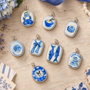 May include: A collection of nine ceramic pendants with silver bails, each featuring blue and white hand-painted designs including a creature, a bowl, a sushi roll, a flower, a ribbon, two fish, a stack of books labeled BOOK, and a rabbit labeled TAMA.