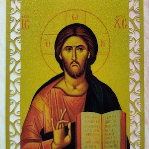 May include: A gold-framed icon of Jesus Christ holding an open book. The book has text in a foreign language. The icon is on a pink background.
