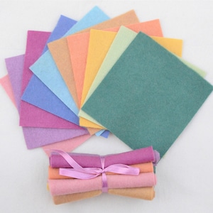 May include: A selection of pastel-colored felt squares arranged in a fan. Below, a bundle of rolled felt in matching hues is tied with a pink ribbon. The felt squares are approximately 20 cm by 20 cm.