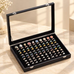 May include: A black rectangular jewelry display case with a clear glass lid and metal latches. The interior features a black velvet lining with multiple rows of slots designed to hold and organize a large collection of various rings.