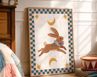 May include: A framed rectangular wall art print featuring a brown rabbit wearing a blue and white ruffled collar with a red bow. The central illustration is surrounded by a pink and teal checkered border and includes yellow stars and crescent moons.