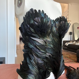 May include: A strapless corset top completely covered in iridescent black feathers that display subtle green and purple metallic highlights. The garment is displayed on a white mannequin torso, showcasing a dense, layered feather texture.