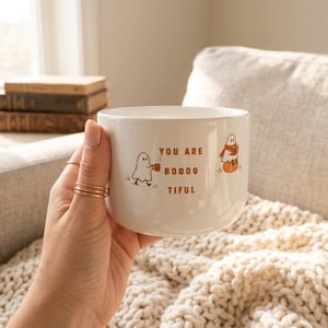 Puede incluir: Una taza de cerámica blanca con un gráfico de dos fantasmas y el texto marrón "YOU ARE BOOOO TIFUL". Un fantasma sostiene una taza, mientras que el otro está sentado sobre una calabaza. La taza tiene una forma redondeada y lisa.