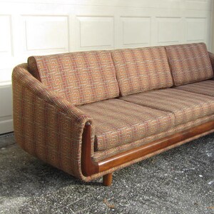 May include: A mid-century modern sofa with a wood frame and a geometric patterned fabric in shades of brown, orange, and yellow. The sofa has three cushions and a slightly curved back.