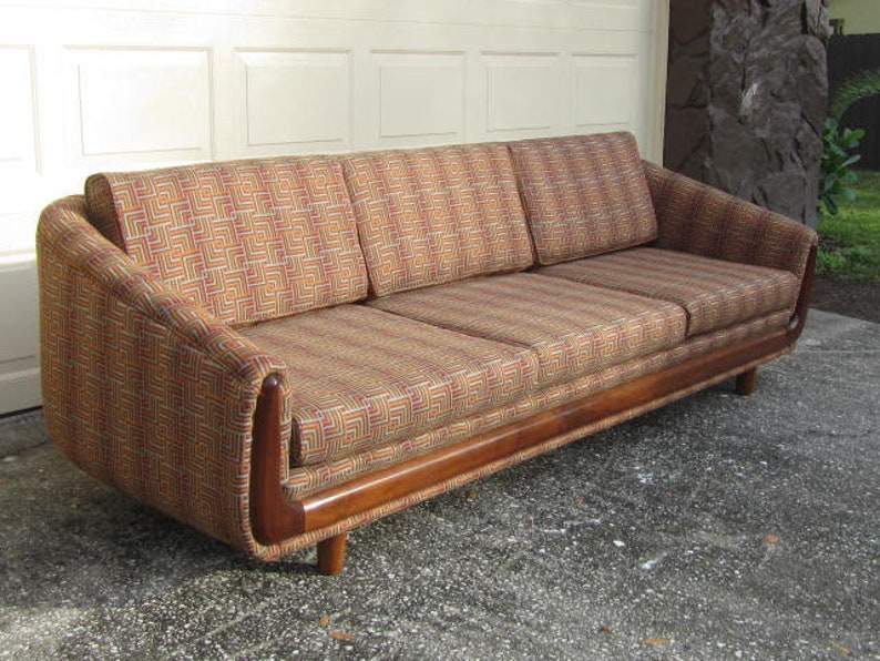 May include: A mid-century modern sofa with a wood frame and a geometric patterned fabric in shades of brown, orange, and yellow. The sofa has three cushions and a slightly curved back.