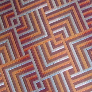 May include: A close-up of a fabric with a geometric pattern of orange, brown, and silver stripes. The fabric has a woven texture.