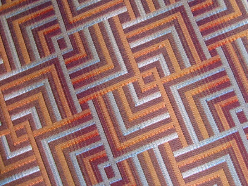 May include: A close-up of a fabric with a geometric pattern of orange, brown, and silver stripes. The fabric has a woven texture.