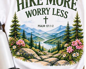 May include: A white short-sleeve t-shirt featuring a graphic of a mountain landscape with a lake, pine trees, and pink flowers. The text reads "HIKE MORE WORRY LESS" with a cross and "PSALM 121:1-2" printed in dark green above the mountain scene.