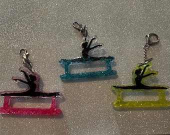 May include: Three acrylic keychains featuring a black silhouette of a gymnast performing a split on a balance beam. The base of each keychain is filled with glitter in pink, blue, or yellow, and each includes a silver-toned metal lobster clasp.