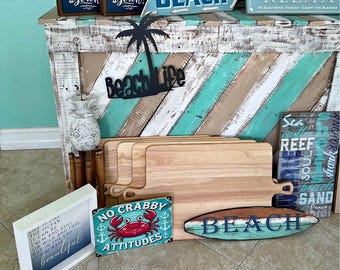 Op de afbeelding: Een verzameling stranddecoraties waaronder houten borden met teksten zoals 'Life is better at the beach', 'Beach Life', 'No Crabby Attitudes' en 'Relax', samen met houten snijplanken, een ananasfiguur en een surfboardvormig bord.