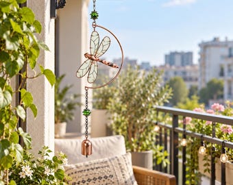 Könnte beinhalten: Ein hängendes Windspiel mit einer kupferfarbenen Libelle mit durchscheinenden Flügeln, die in einem kupfernen Halbmondrahmen zentriert ist. Das Ornament enthält grüne Glasperlen, eine Metallkette und eine kleine Kupferglocke am unteren Ende.