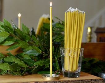 May include: A lit yellow beeswax candle stands on a gold metal lid, next to a glass jar filled with a bundle of unlit yellow beeswax candles. The candles are on a wooden surface with green leaves in the background.