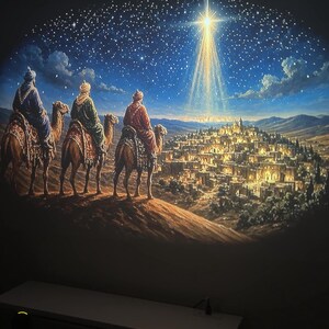 May include: A wall projection displaying a scene of three figures riding camels across a desert landscape towards a glowing city under a night sky. A bright, radiant star shines above the city, casting light over the illuminated buildings.