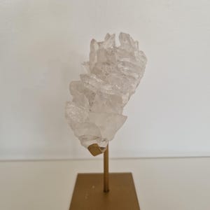 K&ouml;nnte beinhalten: Ein klarer Quarz-Kristallcluster, montiert auf einem goldfarbenen Metallst&auml;nder. Der Kristall hat mehrere Spitzen und eine raue Textur. Der St&auml;nder hat eine quadratische Basis und einen schlanken Stiel.