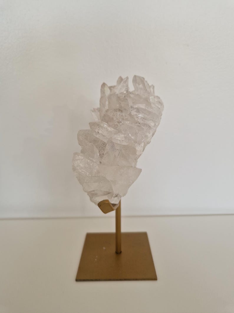 K&ouml;nnte beinhalten: Ein klarer Quarz-Kristallcluster, montiert auf einem goldfarbenen Metallst&auml;nder. Der Kristall hat mehrere Spitzen und eine raue Textur. Der St&auml;nder hat eine quadratische Basis und einen schlanken Stiel.