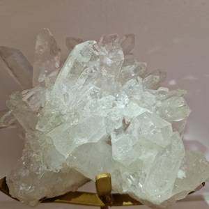 K&ouml;nnte beinhalten: Ein Cluster aus klaren Quarzkristallen, die eine Vielzahl von spitzen Formationen aufweisen. Die Kristalle sind auf einem goldfarbenen Metallst&auml;nder montiert. Der Quarz hat ein durchscheinendes Aussehen und reflektiert das Licht.