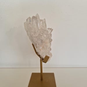 K&ouml;nnte beinhalten: Ein klarer Quarz-Kristallcluster, montiert auf einem goldfarbenen Metallst&auml;nder. Der Kristall hat mehrere Spitzen und ein leicht tr&uuml;bes Aussehen. Der St&auml;nder hat eine quadratische Basis und eine vertikale St&uuml;tze.
