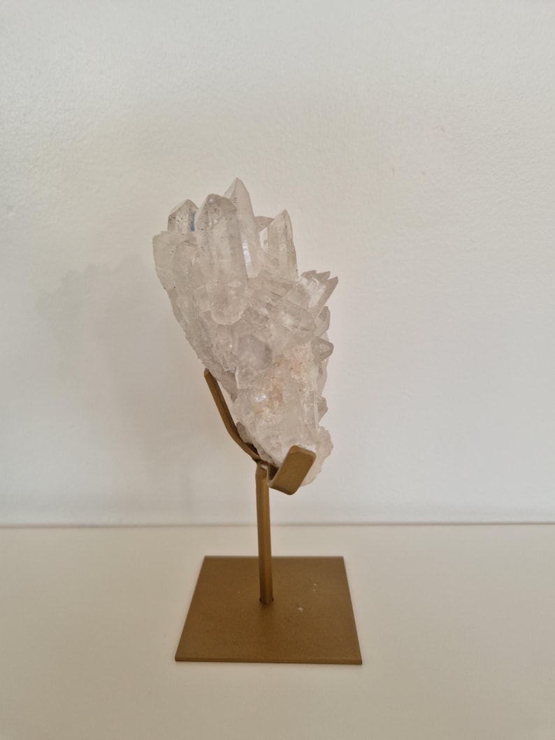 K&ouml;nnte beinhalten: Ein klarer Quarz-Kristallcluster, montiert auf einem goldfarbenen Metallst&auml;nder. Der Kristall hat mehrere Spitzen und ein leicht tr&uuml;bes Aussehen. Der St&auml;nder hat eine quadratische Basis und eine vertikale St&uuml;tze.