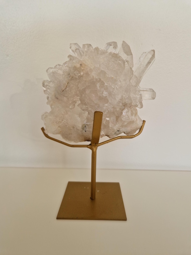 K&ouml;nnte beinhalten: Ein klarer Quarz-Kristallcluster auf einem goldfarbenen Metallst&auml;nder. Der Quarz hat mehrere Spitzen und eine raue Textur. Der St&auml;nder hat eine quadratische Basis und einen schlanken Stiel mit gew&ouml;lbter Oberseite.