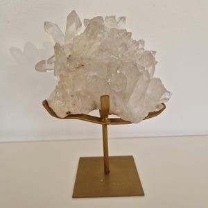 K&ouml;nnte beinhalten: Ein klarer Quarz-Kristallcluster auf einem goldfarbenen Metallst&auml;nder. Der Quarz hat mehrere Spitzen und eine raue Textur. Der St&auml;nder hat eine quadratische Basis und eine gebogene Oberseite, um den Kristall zu halten.
