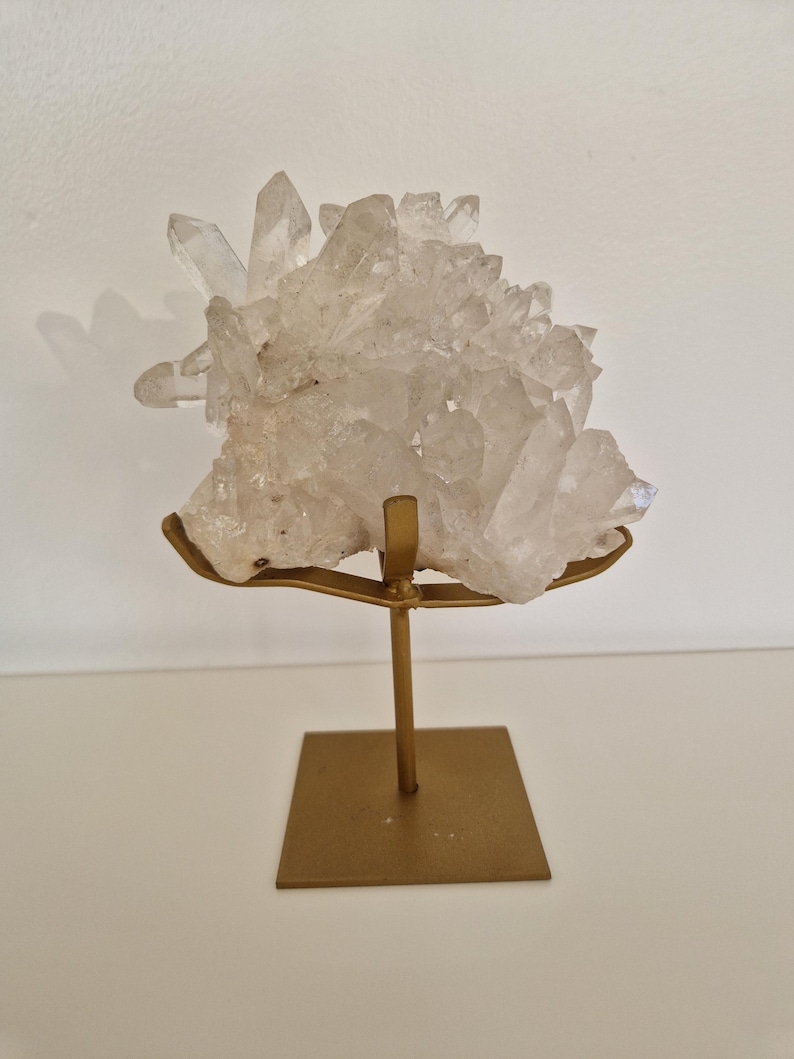 K&ouml;nnte beinhalten: Ein klarer Quarz-Kristallcluster auf einem goldfarbenen Metallst&auml;nder. Der Quarz hat mehrere Spitzen und eine raue Textur. Der St&auml;nder hat eine quadratische Basis und eine gebogene Oberseite, um den Kristall zu halten.