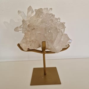 K&ouml;nnte beinhalten: Ein klarer Quarz-Kristallcluster auf einem goldfarbenen Metallst&auml;nder. Der Quarz hat mehrere Spitzen und eine raue Textur. Der St&auml;nder hat eine quadratische Basis und einen schlanken Stiel.
