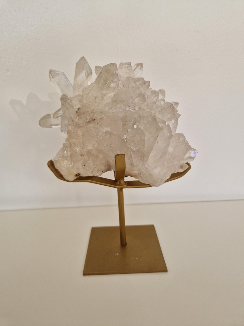 K&ouml;nnte beinhalten: Ein klarer Quarz-Kristallcluster auf einem goldfarbenen Metallst&auml;nder. Der Quarz hat mehrere Spitzen und eine raue Textur. Der St&auml;nder hat eine quadratische Basis und einen schlanken Stiel.
