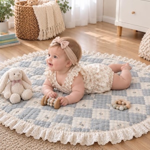 May include: A round, quilted play mat featuring a blue and white checkered pattern with small floral accents and a ruffled lace border. The mat is accompanied by a plush cream-colored rabbit toy and two wooden bead rattles.