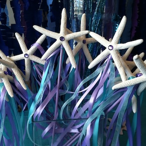 May include: A group of white starfish wands with purple and blue ribbons. The wands are arranged in a vase and are ready to be used for a party or celebration.