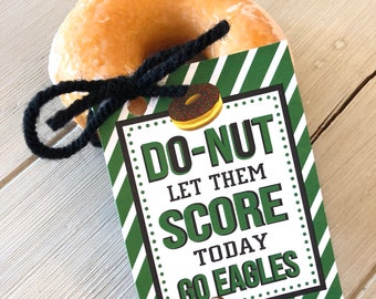 Può includere: Un'etichetta a strisce verdi e bianche con il testo "Do-Nut Let Them Score Today Go Eagles" e un'immagine di un pallone da calcio. L'etichetta è legata a un donut glassato con uno spago nero.