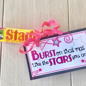 May include: A black card with white text that reads "BURST on that mat Like the STARS you are" with a pink ribbon and a yellow candy bar.