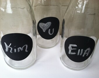 May include: Three clear glass bottles with black chalkboard labels. The labels have white chalk writing on them. The labels say "Kim", "U" with a heart, and "Ella".