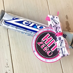 May include: A white chocolate bar wrapped in silver foil with the word "Zero" printed on it. A pink and white ribbon with the text "Go Team" is tied around the bar. A round, black and white tag with the text "Hit Zero" is attached to the ribbon.