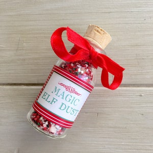 May include: A small glass bottle filled with red and white sprinkles, tied with a red ribbon and a cork top. The bottle has a label that reads "Magic Elf Dust".