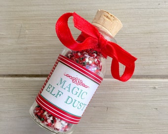 Puede incluir: Una pequeña botella de vidrio llena de chispas rojas y blancas, atada con una cinta roja y un tapón de corcho. La botella tiene una etiqueta que dice "Magic Elf Dust".