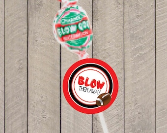 Può includere: Un lecca-lecca avvolto con un design rosso, verde e bianco. Un adesivo circolare con il testo "BLOW THEM AWAY" e una grafica di football è attaccato allo stecco del lecca-lecca. Lo sfondo è un legno chiaro.