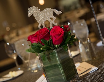 May include: A bouquet of red roses in a clear glass vase with a silver glitter horse figurine on top. The vase is sitting on a table with a grey tablecloth.
