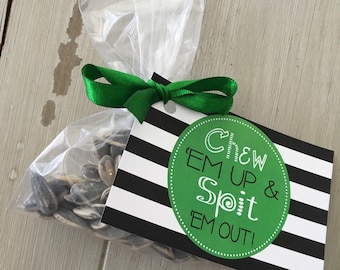 Puede incluir: Una bolsa de plástico transparente llena de semillas de girasol, atada con una cinta verde y una etiqueta a rayas blancas y negras que dice "Chew 'em up & Spit 'em out!"