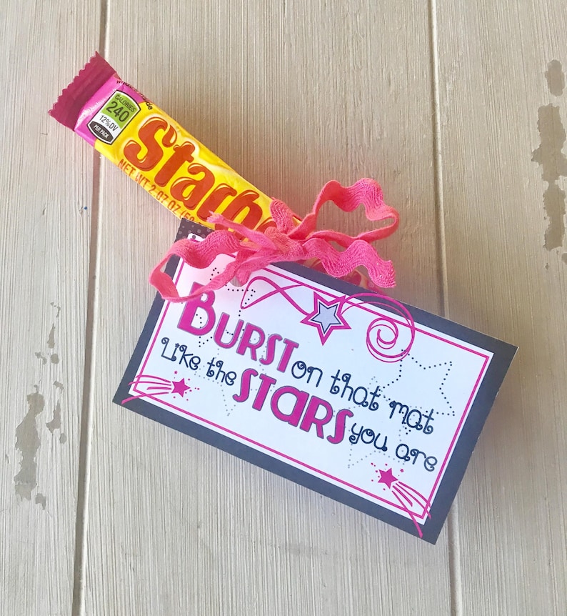 May include: A black card with white text that reads "BURST on that mat Like the STARS you are". The card is decorated with pink and silver stars. A yellow candy bar is tied to the card with a pink ribbon.