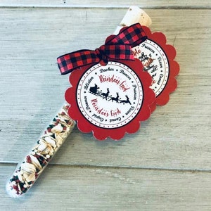 May include: A clear glass tube filled with a mixture of grains and red sprinkles, topped with a cork and a red and black plaid ribbon. Two red scalloped tags with the words "Reindeer Food" and the names of Santa's reindeer are attached.