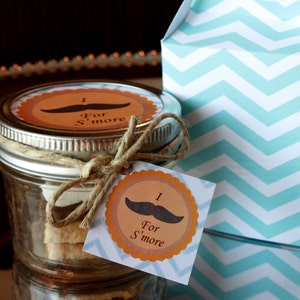 May include: A glass jar filled with layers of graham crackers, chocolate, and marshmallows. The jar is tied with twine and has a brown and orange tag that says "I for S'more".