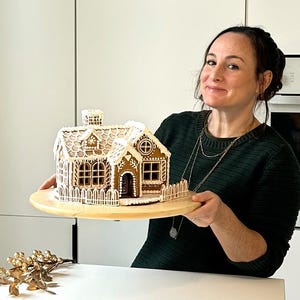 May include: A gingerbread house on a round wooden board, featuring brown walls with white icing details, a chimney, a roof with snowflake patterns, and a white picket fence surrounding the base of the structure.