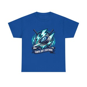May include: Royal blue t-shirt featuring a hockey-themed graphic. The design includes a hockey puck, crossed hockey sticks, and lightning bolts, with the text "TAMPA BAY LIGHTNING" on a banner. The graphic is in shades of blue and white.