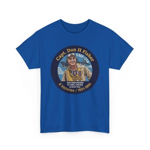 May include: A royal blue t-shirt featuring a circular graphic of Capt. Don H Fisher in flight gear with a plane in the background. The text reads Capt. Don H Fisher, VMF-214, Distinguished Flying Cross w/Gold Star, 6 Victories / 1921-1995.