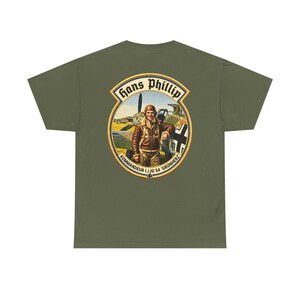 May include: An olive green t-shirt featuring a circular graphic on the back. The design depicts a pilot in a brown leather jacket standing before a fighter plane, with the text "Hans Phillip" and "KOMMANDEUR I./JG 54 'GRÜNHERZ'" printed around it.