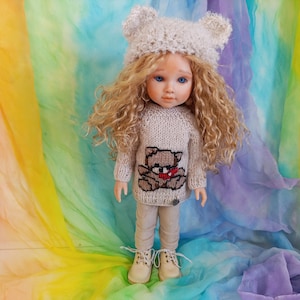 Peut inclure: Une poupée aux longs cheveux blonds bouclés portant un bonnet gris clair duveteux avec des oreilles d'ours, un pull en tricot gris clair à manches longues avec un motif d'ours brun et un cœur rouge, un pantalon beige clair et des chaussures à lacets.